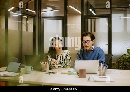 Young biracial male and female colleagues sitting at table in modern office looking at laptop screen and discussing work Stock Photo
