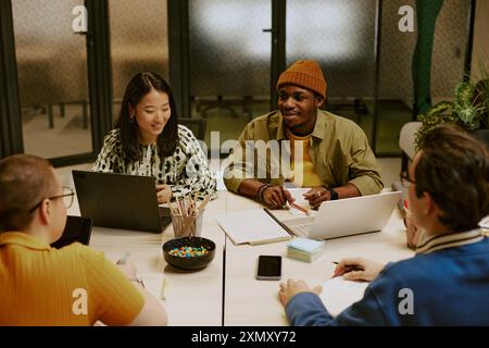 High angle view of multi-ethnic group of young colleagues sitting at table with laptops, gadgets and documents on top, discussing work plan Stock Photo