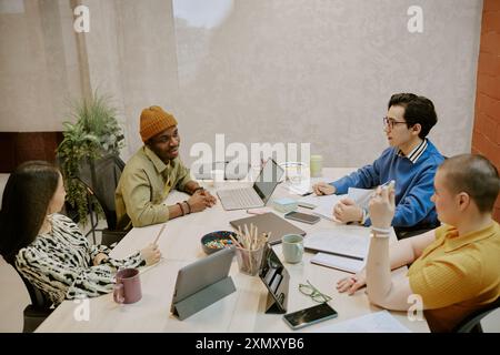 High angle view of multi-ethnic gen Z managers discussing plan while sitting at table in modern office equipped with laptops and gadgets Stock Photo