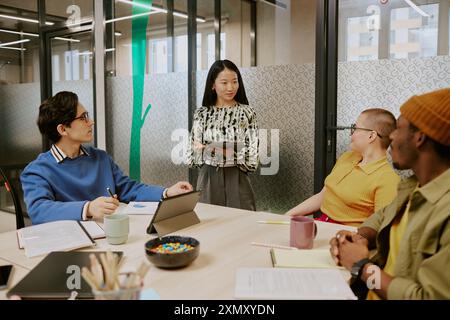 Young Asian manager holding digital tablet standing in front of her multi-ethnic colleagues speaking about project during meeting Stock Photo