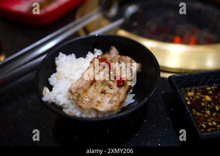 Bowl of white rice topped with grilled meat and red chili slices, paired with dipping sauce on a black stone tabletop. Stock Photo