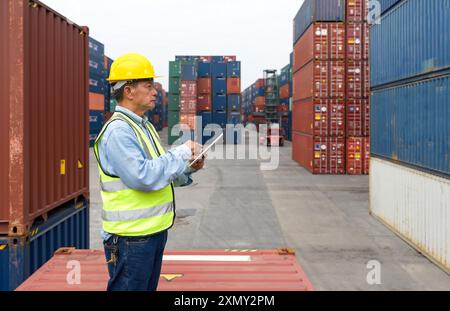 A worker inspects shipping containers in a busy port, wearing a safety vest and hard hat, holding tablet computer. Stock Photo