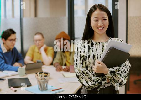 Medium portrait of cheerful young Asian female specialist holding documents and digital tablet standing in office and smiling at camera Stock Photo