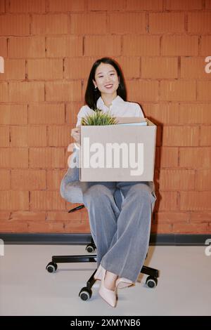 Vertical long shot of cheerful young Asian female new employee sitting on chair with box on lap against red brick wall Stock Photo