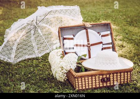 Romantic set with vintage picnic basket, lace sun umbrella and summer hat on park garden lawn on sunny summer day outdoors. Stock Photo