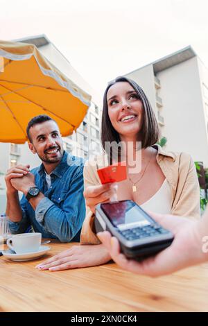 Vertical couple paying the bill of the coffee shop using a contactless creditcard payment. Young adult customer woman doing a purchase on a restaurant Stock Photo