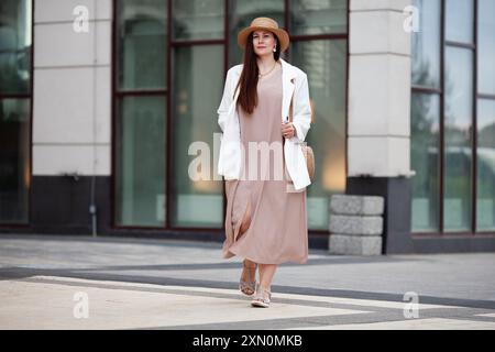 Stylish Plus size woman walks confidently outdoors in urban city, wearing beige dress, white blazer, wide brimmed hat, woven handbag. Model with long Stock Photo