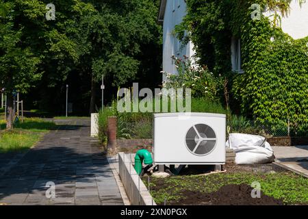 A technician works on installing an air conditioning unit in a lush residential garden, surrounded by greenery on a bright, sunny afternoon. Stock Photo