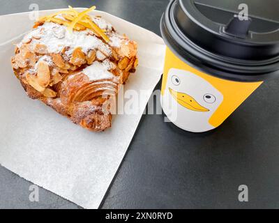 Takeaway coffee in yellow cardboard cup with duck and almond croissant in paper on table in cafe. Stock Photo