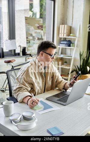 A young man with Down syndrome sits at a desk working on a laptop and phone. Stock Photo