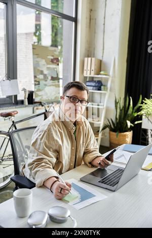 A young man with Down syndrome works on his laptop and a sticky note, looking focused and determined. Stock Photo