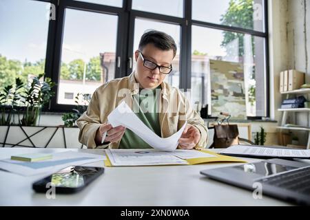 A young man with Down syndrome, dressed in a tan jacket and green shirt, reads papers at a desk. Stock Photo