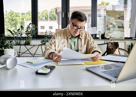 A young man with Down syndrome sits at his desk, reviewing documents while wearing glasses. Stock Photo