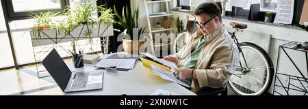 A young man with Down syndrome sits at a desk reviewing documents, while a bicycle rests against the wall behind him. Stock Photo