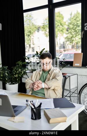 A young man with Down syndrome checks his watch while seated at a desk in an office. Stock Photo