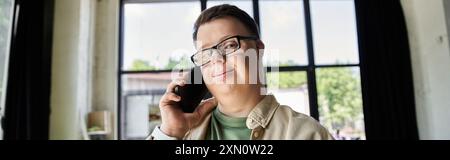 A young man with Down syndrome speaks on a mobile phone while standing near a window. Stock Photo