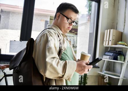 A young man with Down syndrome, wearing glasses, checks his phone while holding a cup of coffee. Stock Photo