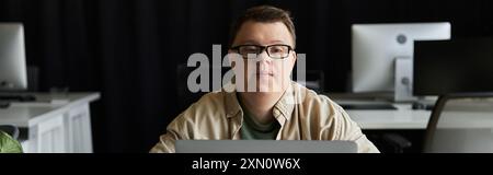 A young man with Down syndrome sits at a desk in an office, working on his laptop. Stock Photo