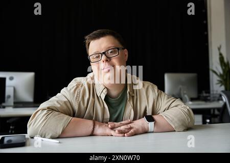 A young man with Down syndrome sits at a desk with a relaxed, confident smile. Stock Photo