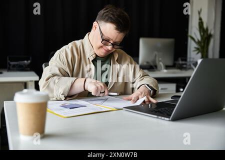 Young man with Down syndrome works hard at desk, laptop and coffee nearby Stock Photo