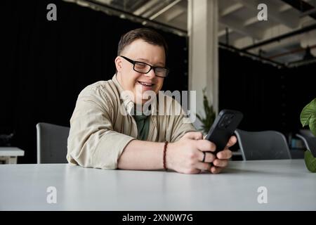 A young man with Down syndrome smiles while using his phone at a table. Stock Photo