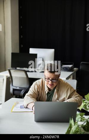 A young man with Down syndrome sits at a desk, working on a laptop. Stock Photo