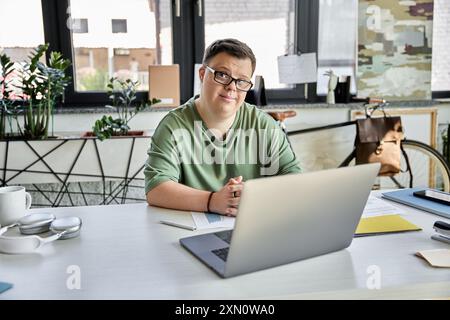 A young man with Down syndrome sits at a desk, smiling while using his laptop. Stock Photo