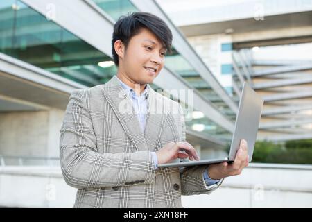 Young Asian Man Using Laptop Outside a Modern Building Stock Photo
