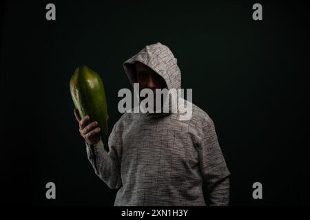 Mysterious man with glasses wearing hoodie holding green papaya. Isolated on dark background. Stock Photo