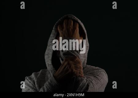 Close up portrait of mysterious man wearing sunglasses and hooded sweatshirt covering head with hands on face. Isolated on darkened background. Stock Photo