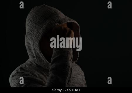 Close up portrait of mysterious man wearing sunglasses and hooded sweatshirt covering head with hands on face. Isolated on darkened background. Stock Photo
