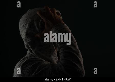 Close up portrait of mysterious man wearing sunglasses and hooded sweatshirt covering head with hands on face. Isolated on darkened background. Stock Photo