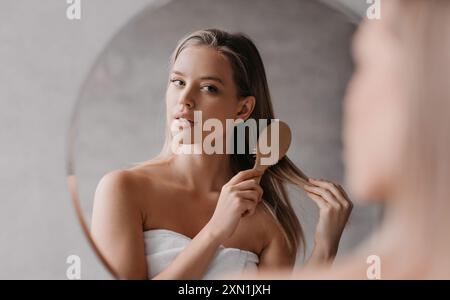 Young cute lady wrapped in towel, standing in front of mirror at bathroom and combing her hair after shower Stock Photo