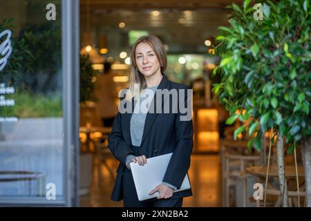 Confident entrepreneur woman stands outside contemporary cafe holds laptop, female restaurant owner Stock Photo