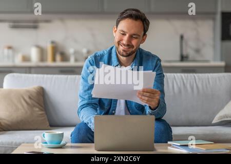 Smiling Man Reviewing Documents While Using Laptop on Sofa Stock Photo