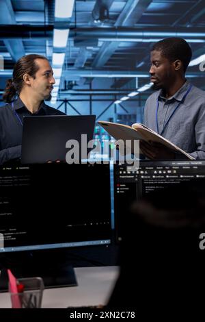 Manager getting feedback from engineer while overseeing data center, looking for firewall misconfigurations. Supervisor talking with workers in server room, asking about systems bottlenecks Stock Photo