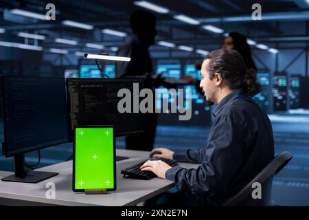Programmer works on green screen tablet in data center housing storage equipment. IT professional using mockup device in server farm with networking components providing vast amounts of computing resources Stock Photo