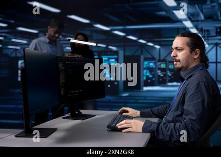 Programmer typing on PC in data center, updating systems to protect servers from exploitable software vulnerabilities. IT expert coding and looking for errors causing equipment to slow down Stock Photo