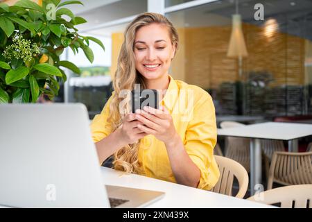 Woman using smartphone at outdoor cafe, female freelancer person in yellow shirt sitting at street restaurant terrace with cellphone looking at device screen and smile. Freelance remote work concept. Stock Photo