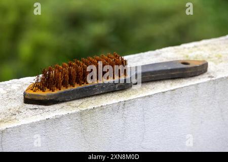 Old wire brush with bristles that are starting to break and rusty is placed on white wall, blurred green nature background Stock Photo