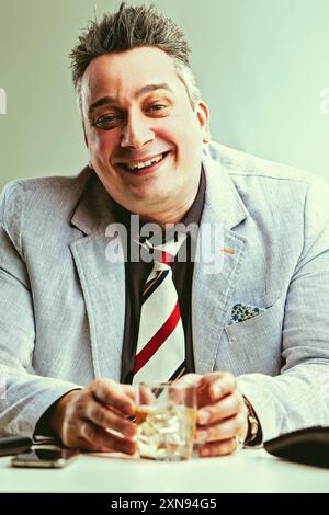 Confident businessman taking a break at his office table, smiling and enjoying a glass of whiskey Stock Photo