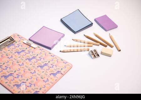 A pink clipboard with a unicorn design sits on a white table. On the clipboard, there are several colorful sticky notes and a pencil. The pencils are Stock Photo