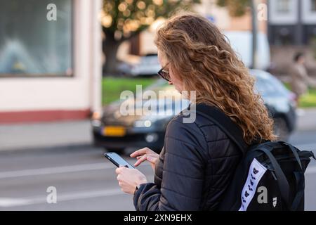 Riga, Latvia - September 30, 2020: A woman waits at a bus stop, focused on her phone. Stock Photo