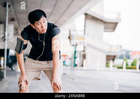 Tired Asian guy in headphones leaning on knees and looking away while listening to music and relaxing during outdoor workout on city street Stock Photo