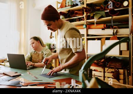 Male tailor preparing leather workpieces while his young female intern using laptop in background Stock Photo