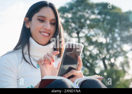 Young Latin Woman sitting in park is using phone touching screen with her finger while reading book, she is happy smiling, there is tree behind her an Stock Photo