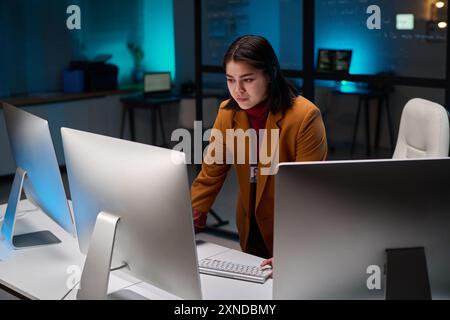 Portrait of female cybersecurity expert using computer with multiple monitors in office lit by blue light Stock Photo