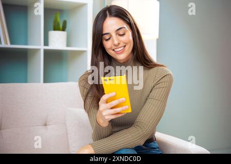 Smiling young woman sitting on a sofa looking at social media with a smartphone Stock Photo