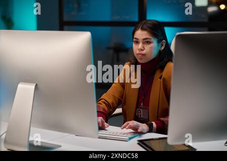 Portrait of successful young woman as female cybersecurity expert using computer in office and looking at screen with blue light Stock Photo