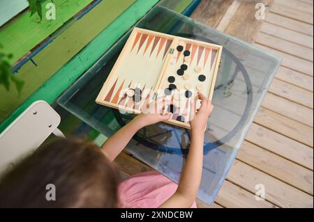 A child engaging in a game of backgammon at a glass table on a vibrant, green and wooden deck, showcasing leisure and strategic thinking. Stock Photo